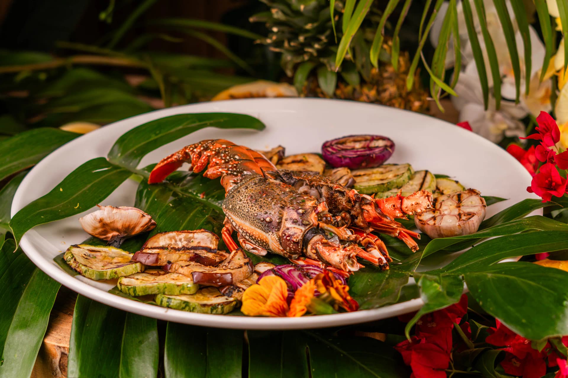 Langosta grillada con vegetales en plato blanco sobre hojas tropicales
