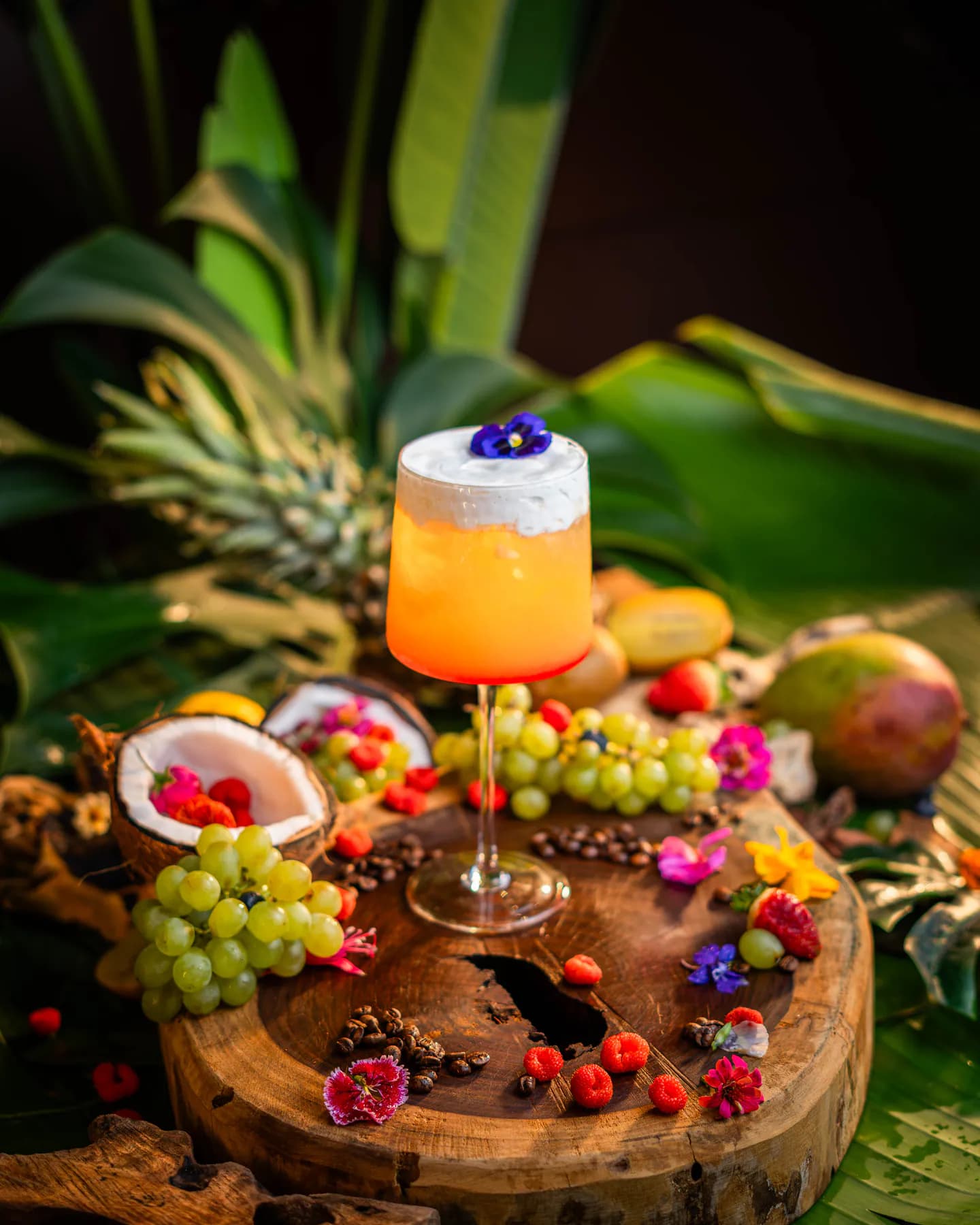 Trago naranja con espuma y una flor azul en copa, sobre un tronco con coco y uvas